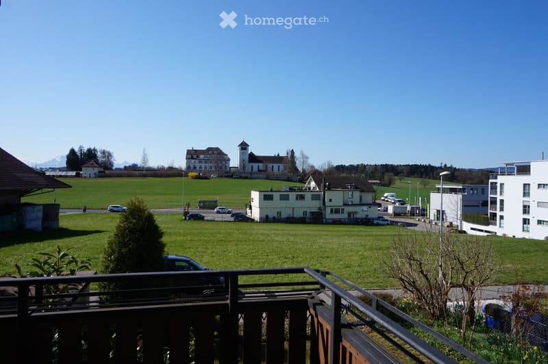 Balcony overlooking a green field with a church, houses, and parked cars in the background.