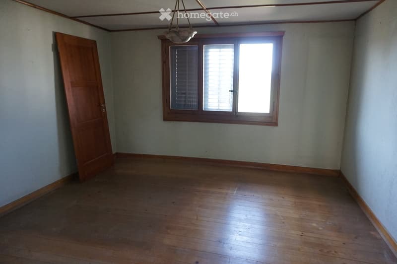Empty room with wooden flooring, light-colored walls, a single window with blinds, and a wooden door.