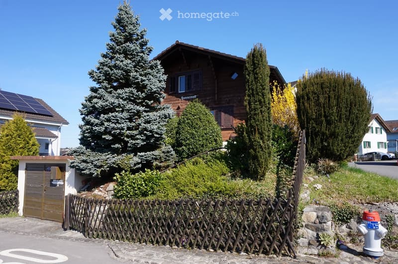 A two-story wooden house with solar panels on the roof, surrounded by tall evergreen trees and shrubs behind a wooden fence.