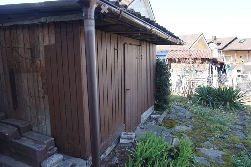 A small wooden shed with a covered entrance, surrounded by a garden area with plants and stepping stones leading to the house.