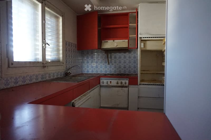 A small kitchen with red cabinets, a sink, a stove, and an oven. The walls are tiled in blue and white patterns, and there is a window with blinds on the left side.