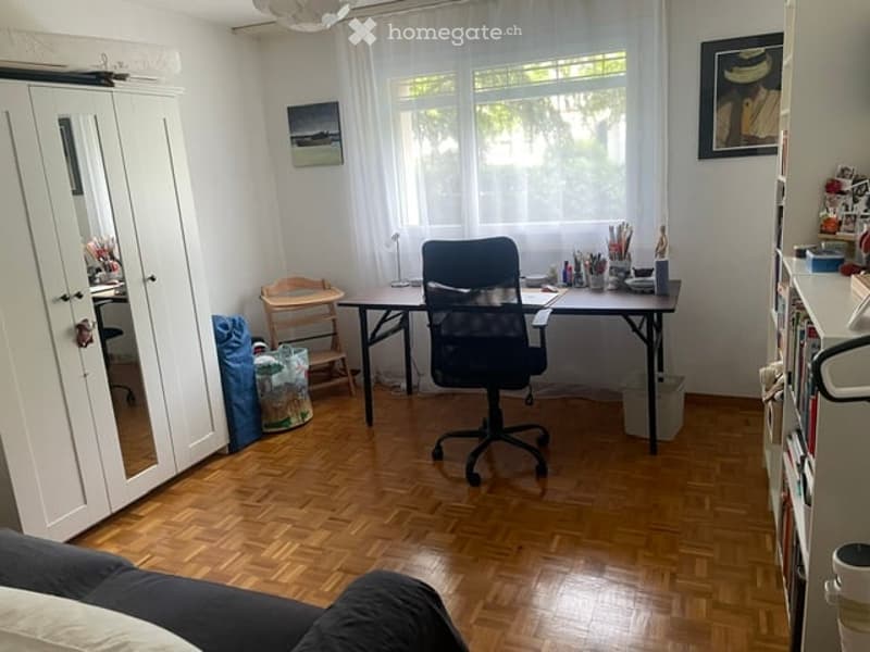 A well-lit room with wooden flooring, a large window with white curtains, a desk with a chair, a small wooden stool, a white cabinet with mirrored doors, and a bookshelf filled with books and decorative items.