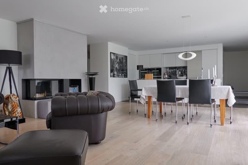 Modern open-plan living and dining area with a dark leather sofa, ottoman, dining table with chairs, fireplace, and kitchen area with cabinets and island.