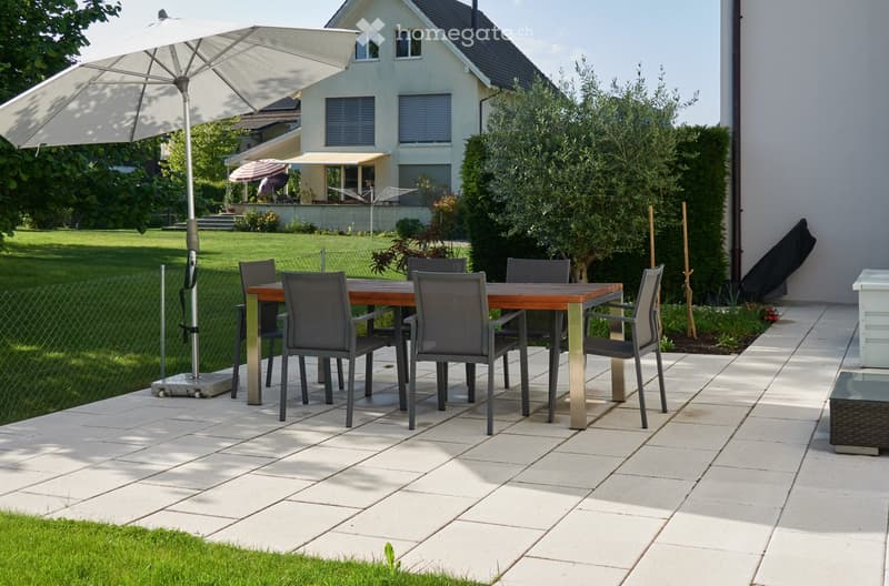 Outdoor patio with a wooden table, six chairs, and a large umbrella, surrounded by a well-maintained lawn and a modern house in the background.