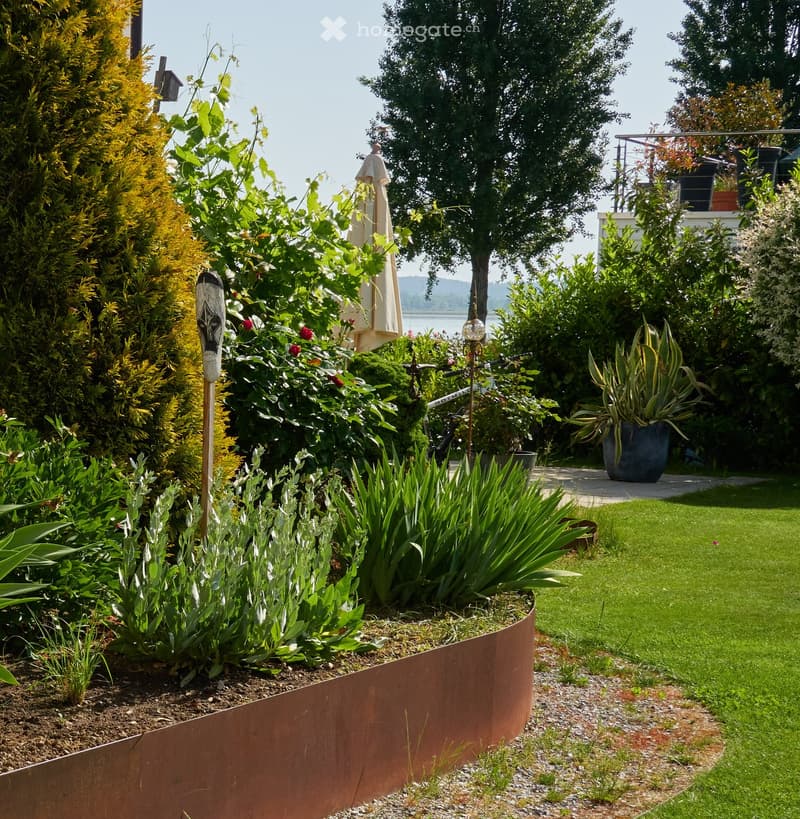 Well-maintained garden with a variety of plants, shrubs, and flowers, including a prominent display of irises and other flowering plants. The garden features a curved metal border, a neatly trimmed lawn, and a pathway leading to a patio area with outdoor
