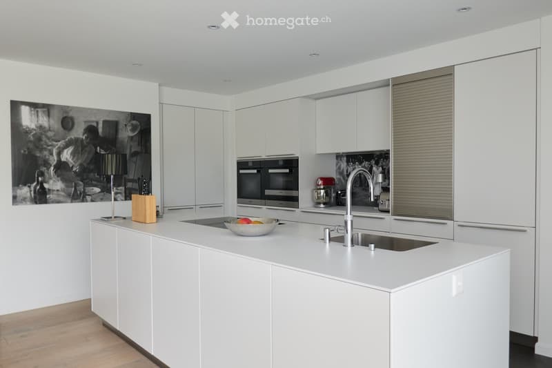 Modern white kitchen with a central island, double sink, built-in ovens, and a mixer. The island features a bowl of fruit and a knife block. The walls are white with a large black-and-white photograph and a lamp on the counter.