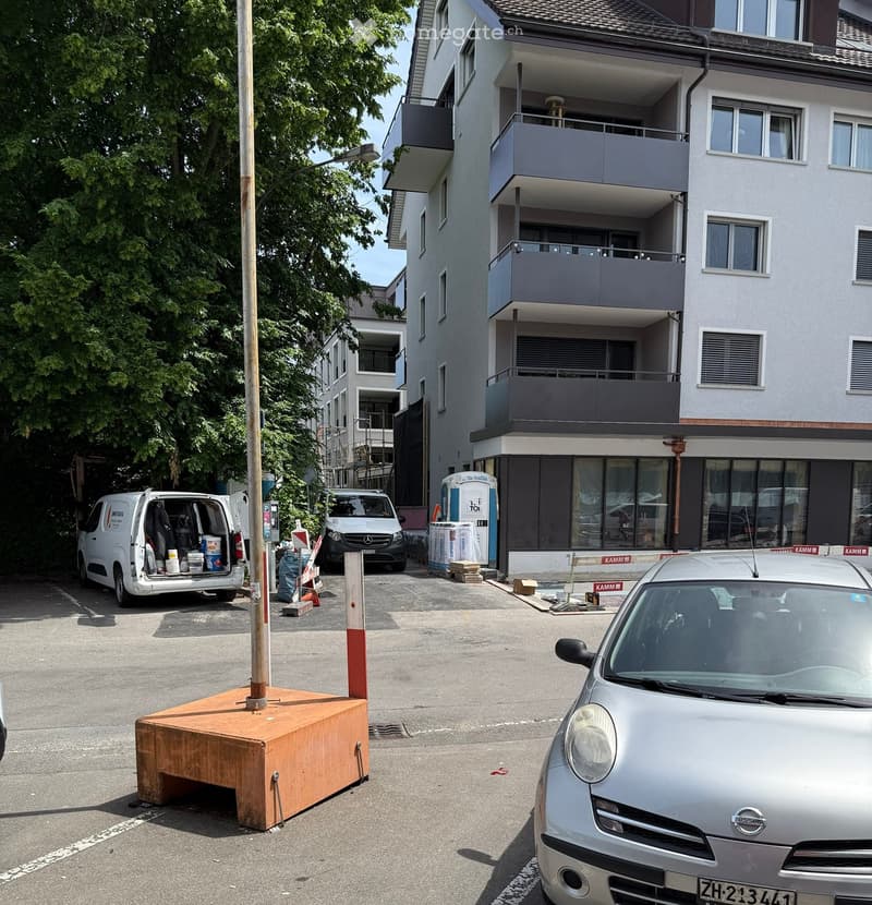 Multi-story residential building with balconies, surrounded by parked cars and construction materials.