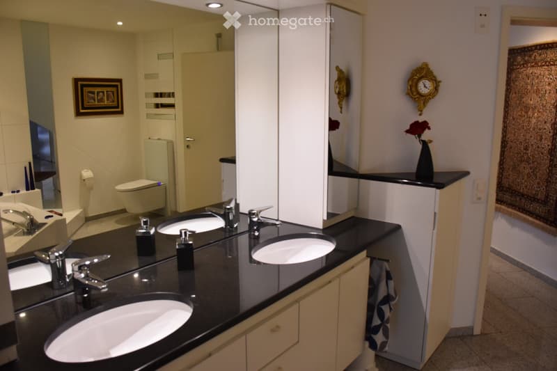Modern bathroom with double sinks, large mirror, toilet, decorative vase, framed picture, and clock on the wall.
