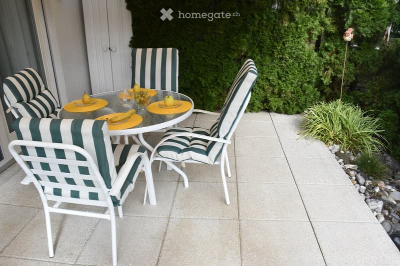 A patio area with a round glass table and four white chairs with green and white striped cushions. The table has a yellow center and holds a bowl of fruit, including grapes, pears, and bananas. The patio is bordered by a low concrete edge with small rocks
