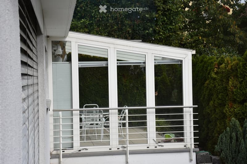 Balcony with white furniture, glass walls, and a metal railing overlooking a green garden.