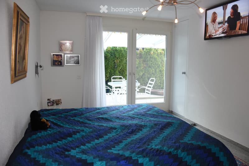 A well-lit bedroom with a large bed covered in a blue and black quilt, a small black dog and a toy on the bed, framed pictures on the wall, a mounted television, and a sliding glass door leading to a patio with outdoor furniture.