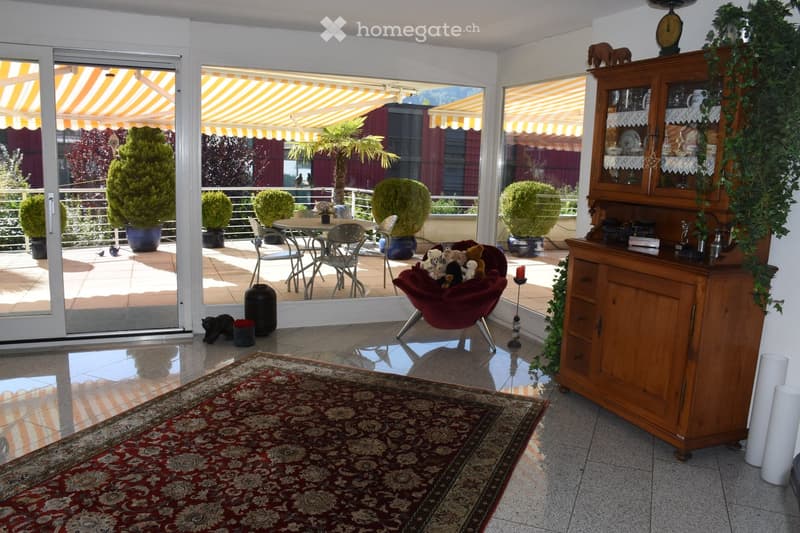 Bright living room with large glass doors leading to a balcony, featuring a red sofa with stuffed animals, a wooden cabinet, a patterned rug, and decorative vases.