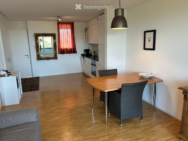 A modern, open-plan living and dining area with a grey sofa, a wooden dining table with two chairs, a kitchen area with cabinets, an oven, and a refrigerator, and a large mirror on the wall.