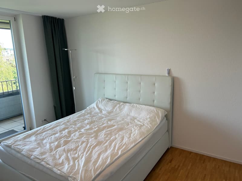 Bedroom with a white upholstered headboard and a neatly made bed, wooden flooring, and a window with dark curtains.