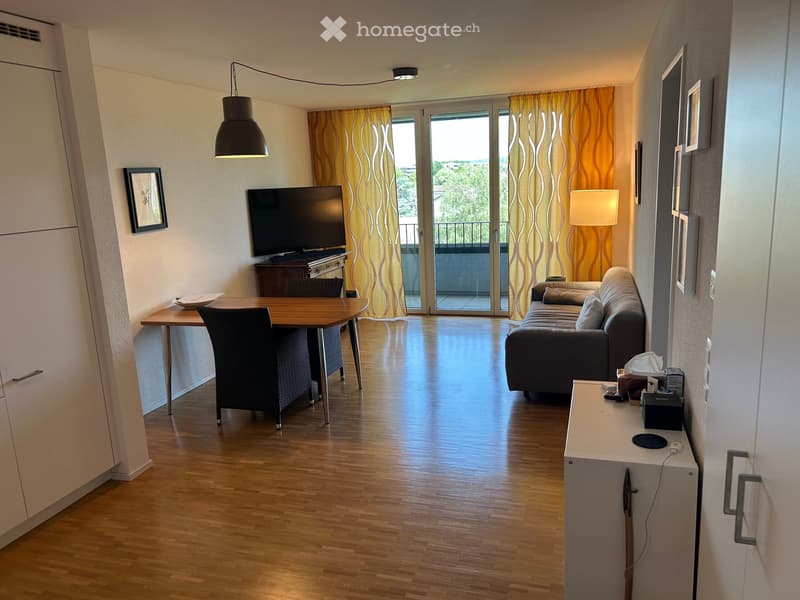 Modern living room with wooden flooring, a sofa, a coffee table, a dining table with chairs, a television, and a lamp. Large windows with yellow curtains lead to a balcony.