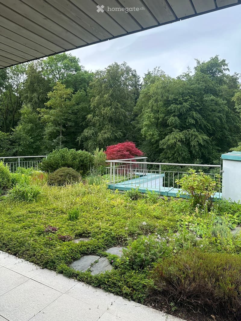 A landscaped garden area with various plants, shrubs, and a small raised platform with a railing, surrounded by trees and greenery.