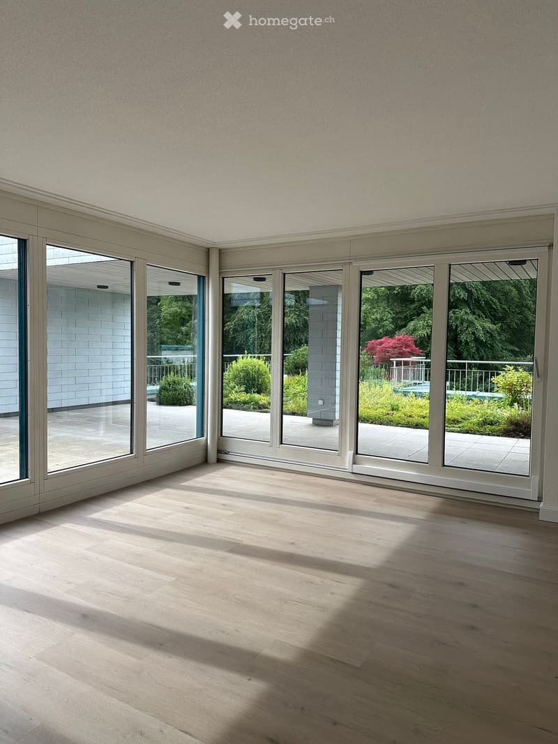 Bright, empty room with light wooden flooring and large sliding glass doors leading to a garden area.