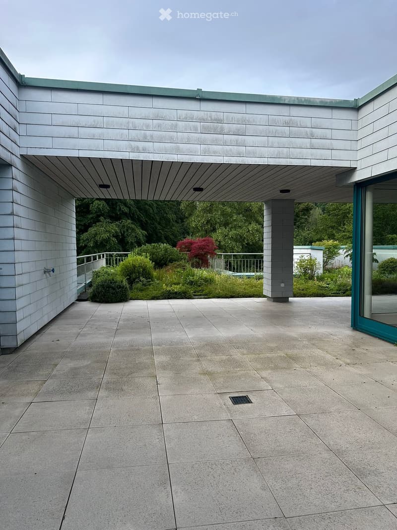 Modern outdoor patio with large concrete tiles, covered walkway, greenery, and a view of trees and shrubs.