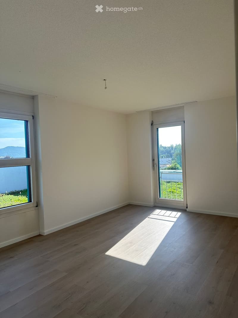 Empty room with light-colored walls, wooden flooring, and large windows offering a scenic view.