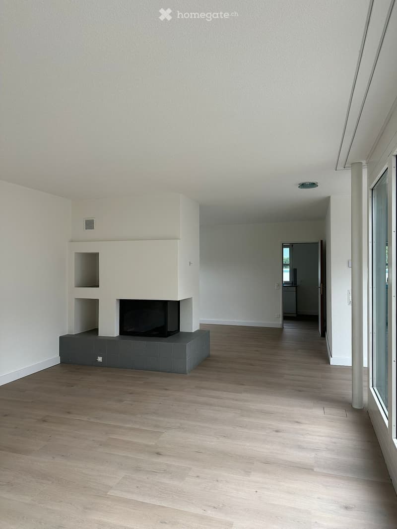 Empty hallway with light wooden flooring, white walls, and a built-in fireplace with a dark tiled base. A doorway leads to another room.