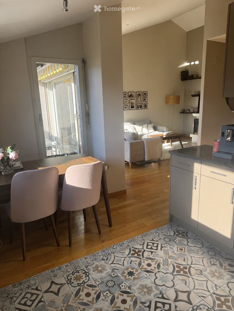 Bright, open-plan living and dining area with wooden flooring, modern furniture, and a view through a large glass door to a balcony. Features include a sofa, coffee table, dining table with chairs, a floor lamp, and a small kitchen area with cabinets and