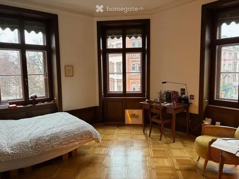 Cozy bedroom with wooden flooring, large windows, a bed, a desk with a chair, a lamp, and a small seating area with a wicker chair.