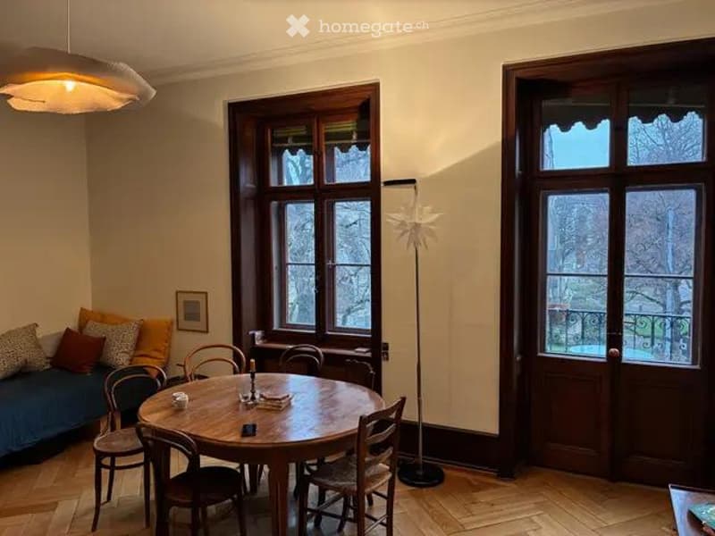 Bright, cozy room with wooden flooring, large windows, a round wooden dining table with chairs, a sofa with colorful cushions, and a floor lamp with a decorative shade.