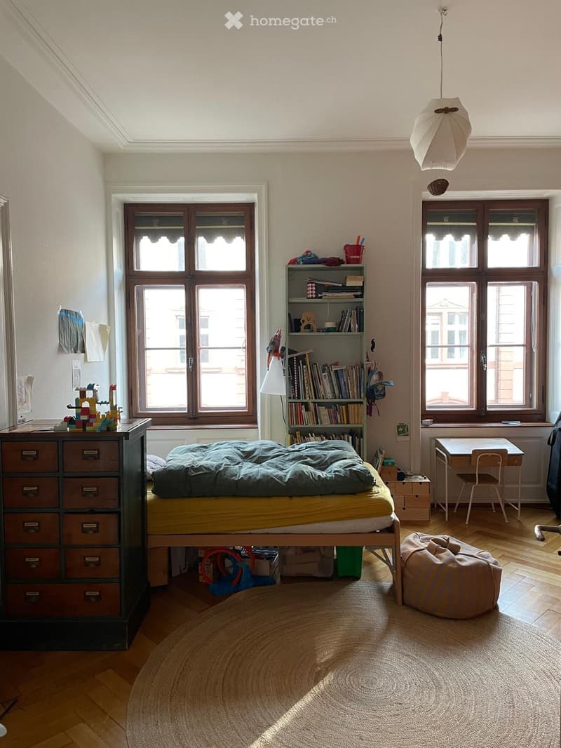 Bright bedroom with wooden floors, large windows, a bed with a green duvet, a bookshelf, a small table with a chair, a bean bag, and a drawer unit.