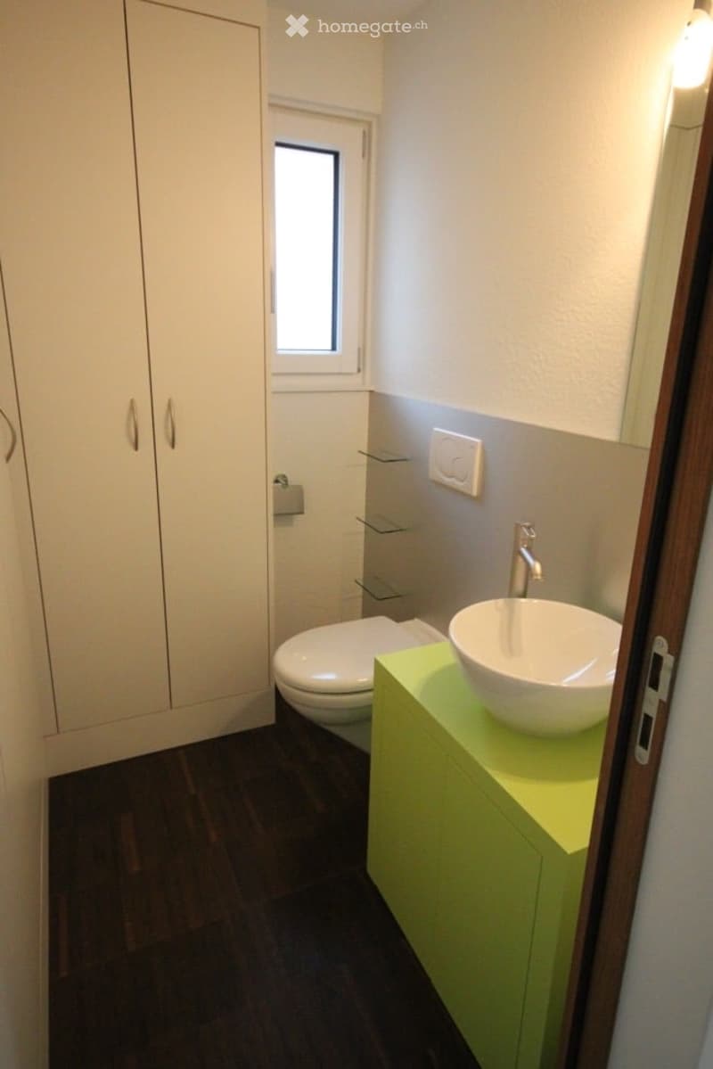Modern bathroom with a white sink, chrome faucet, toilet, and built-in cabinet. Features a small window, glass shelves, and a sleek design.