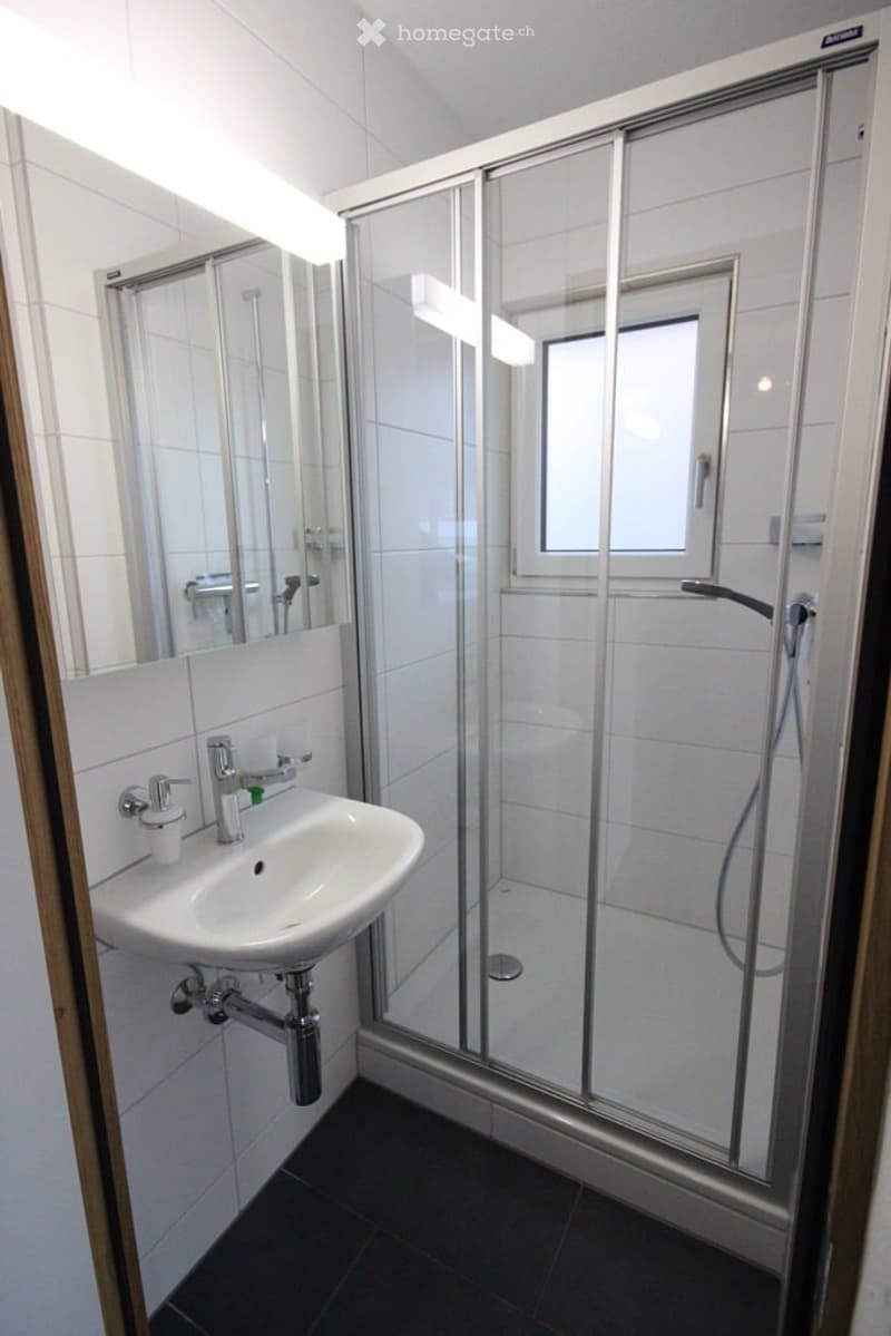 Modern bathroom with white tiles, a glass-enclosed shower, a sink with a mirror, and a window providing natural light.