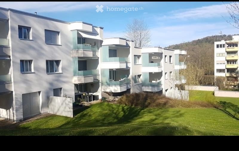 Modern multi-story residential building with white and light green facade, multiple balconies, and a well-maintained lawn in front.