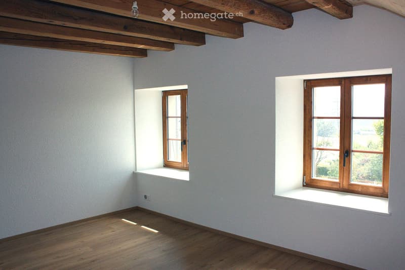 Empty room with wooden flooring, white walls, and two windows with wooden frames. The room features exposed wooden beams on the ceiling and a small built-in shelf beneath one window.