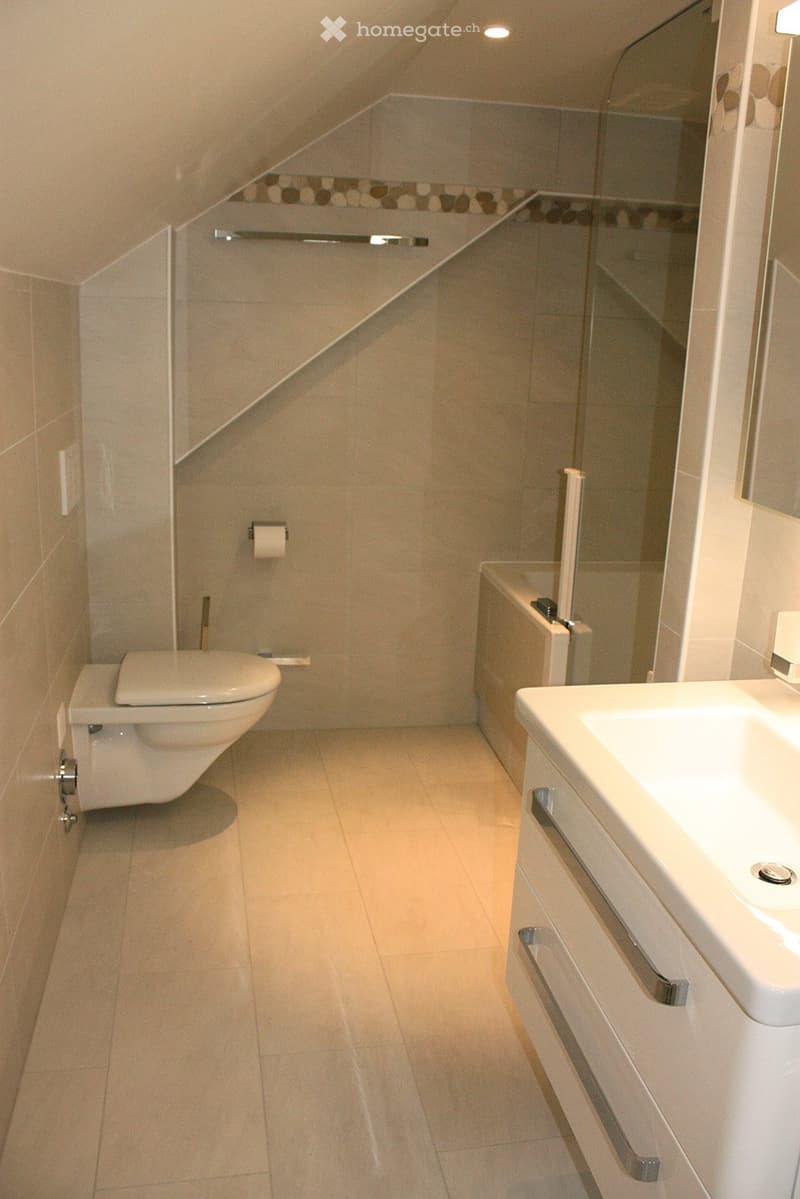 Modern bathroom with a toilet, sink, bathtub, and mirror. The space features light-colored tiles and a sleek design.