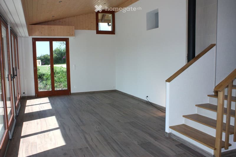 Empty room with wooden flooring, white walls, a staircase leading upwards, and large glass doors opening to a garden.