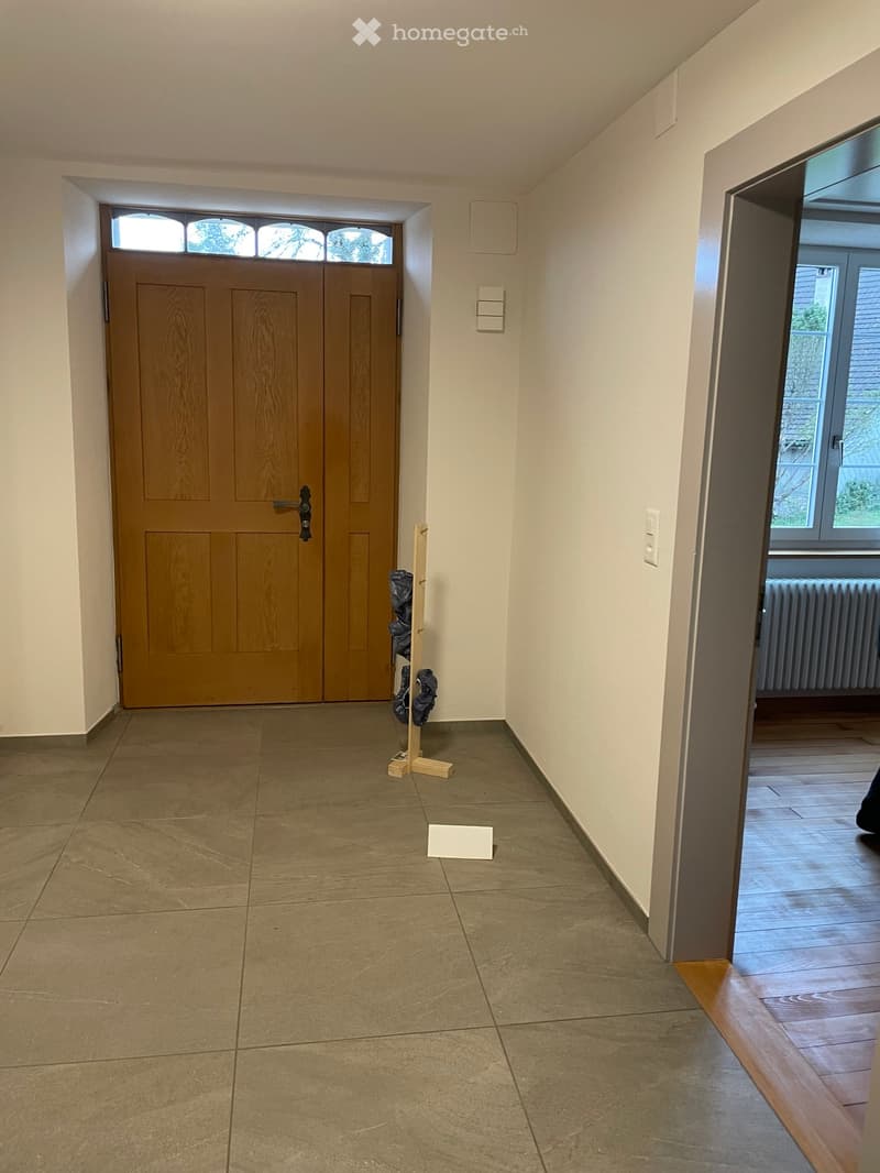Empty hallway with tiled floor, wooden front door, coat rack, and an open doorway leading to another room with wooden flooring and a radiator.