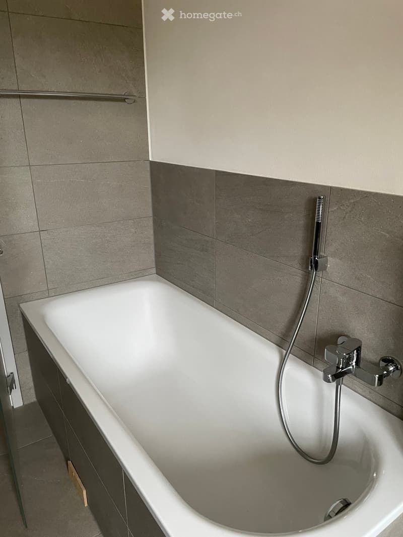 Modern bathroom with a white bathtub, grey tiled walls, and a chrome faucet with a handheld showerhead.