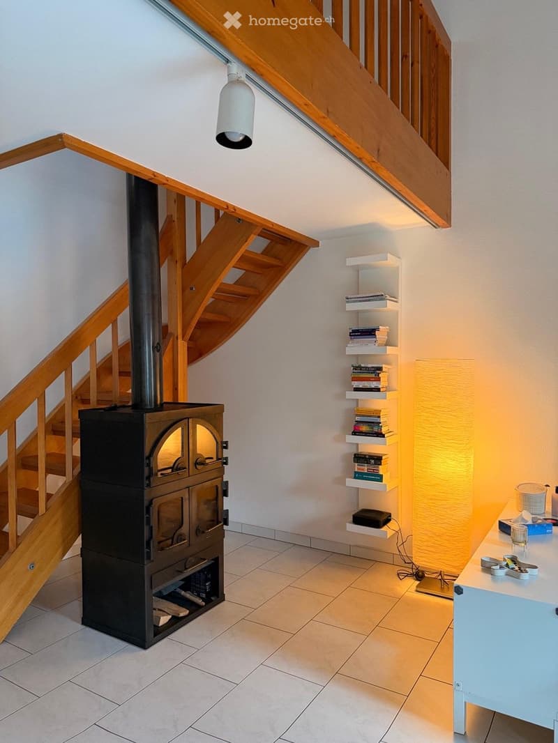 A cozy hallway with a wooden staircase, a black wood-burning stove, a wooden shelf with books, a small table with a lamp, and a hanging light fixture.