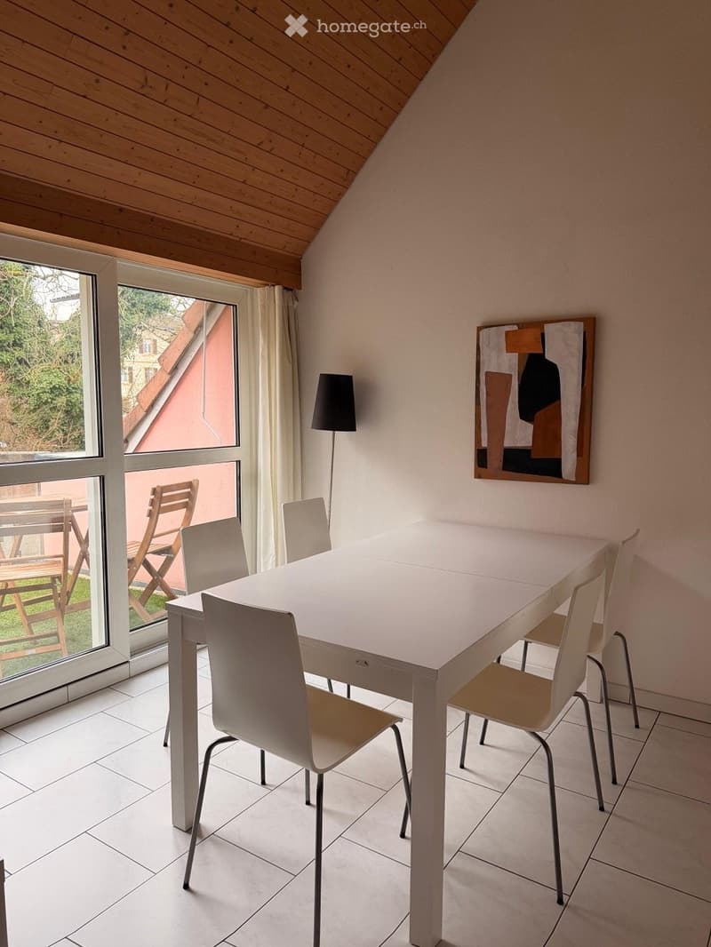 Bright dining area with a white dining table, six chairs, a floor lamp, and a modern painting on the wall. Large windows offer a view of the garden.