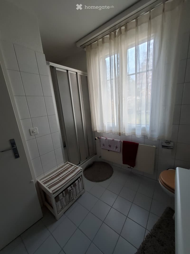 Bathroom with white tiled walls and floor, a toilet, a towel rack with towels, a small storage unit, and a round bathroom rug.