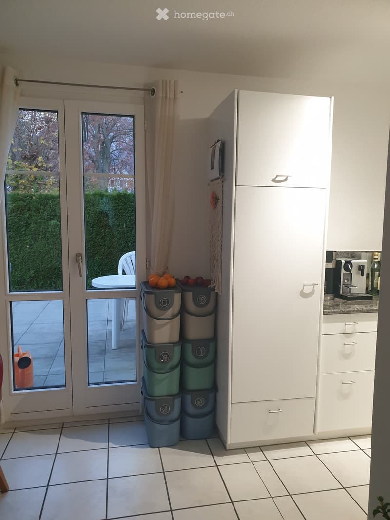 A modern kitchen with white cabinets, a refrigerator, a coffee maker, and a stack of bins. The room features tiled flooring and large windows with a view of the garden.