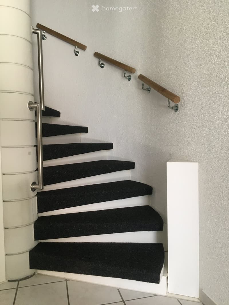 Staircase with black carpeted steps and white risers, featuring a wooden handrail and metal support rail.