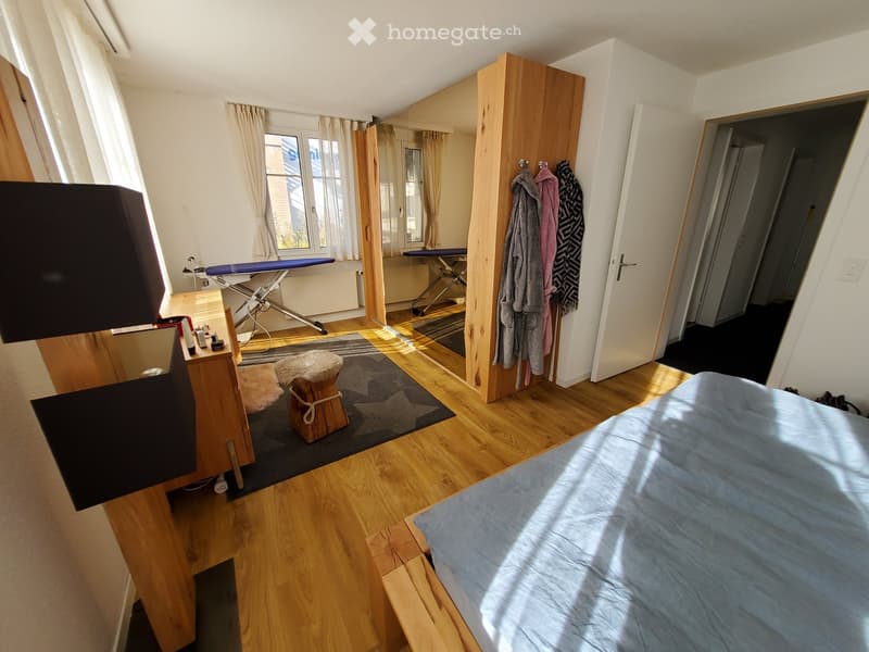 Bright bedroom with wooden flooring, a bed with blue bedding, a wooden wardrobe, a mirror, a small stool, and an ironing board in the corner.