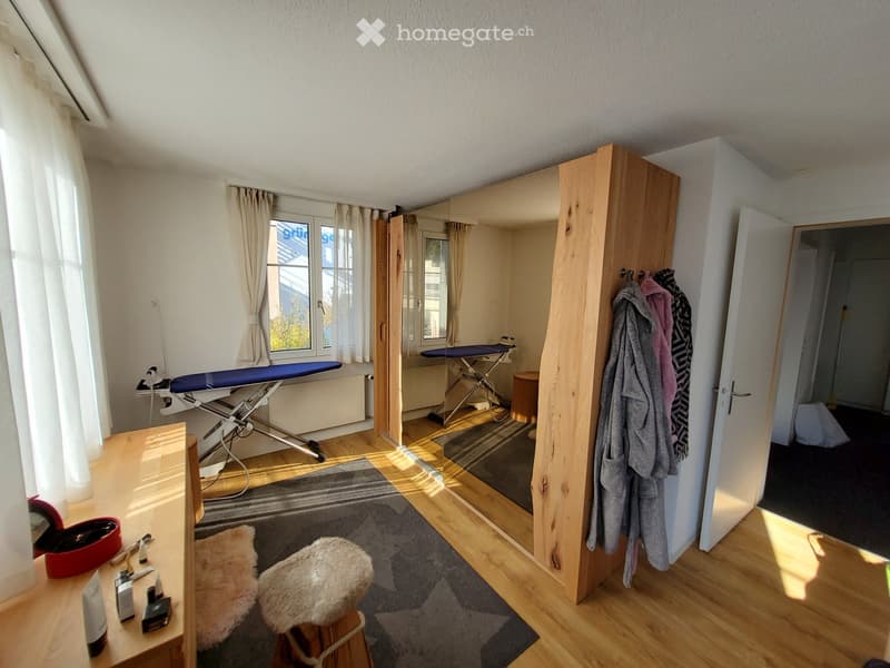 Well-lit room with wooden flooring, large windows, and a variety of furniture including a stool, mirror, ironing board, and a small table with personal care items.