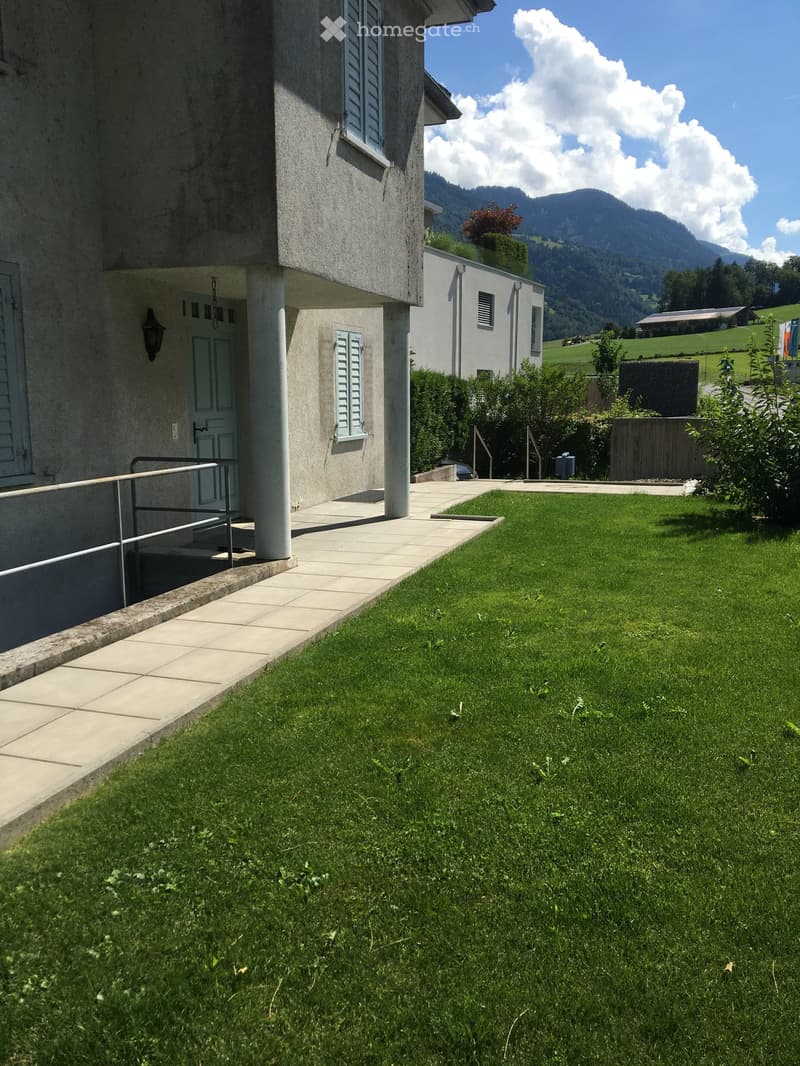 Well-maintained lawn with a concrete pathway leading to a house entrance, surrounded by greenery and distant mountains.