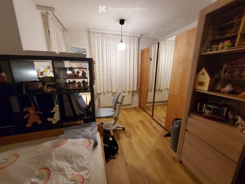 A small, well-lit bedroom with a bed, a chair, shelves filled with toys and books, and a window with curtains.
