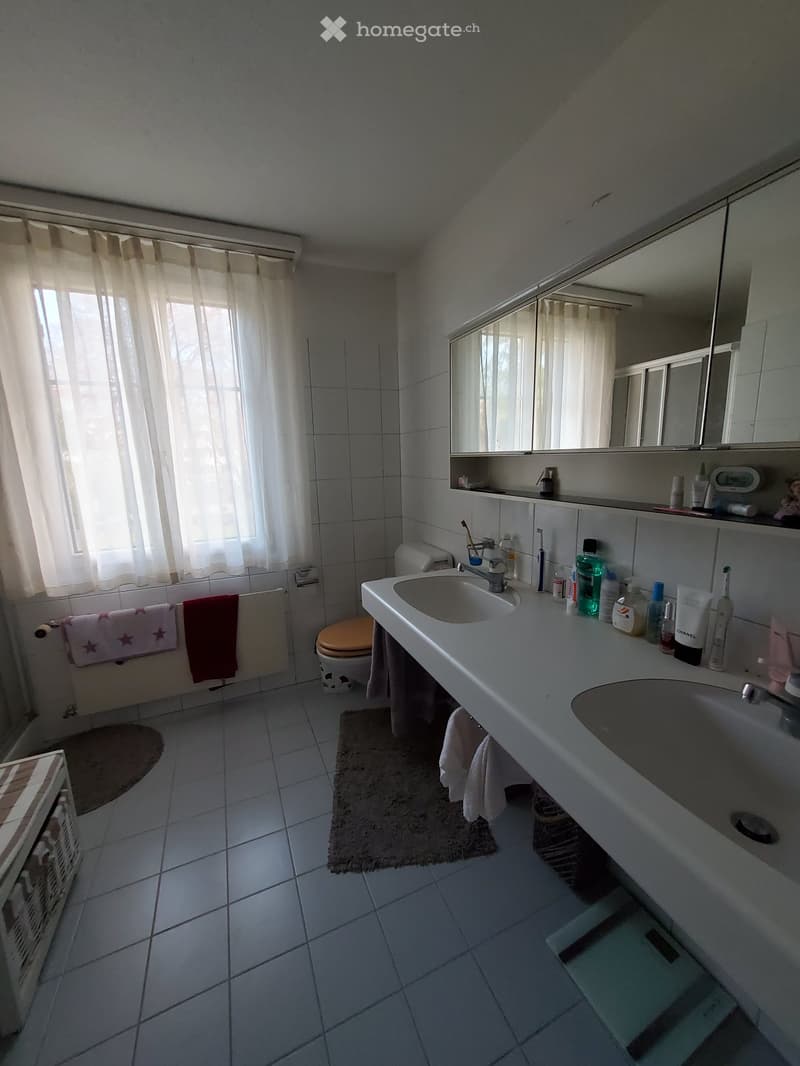 Bathroom with white tiles, a sink, toilet, mirror, and various toiletries on the counter. Towels are hanging on the wall, and a small rug is placed on the floor.