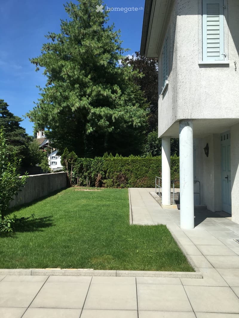 Well-maintained lawn with trimmed hedges, a concrete pathway leading to a house entrance with white columns and a small porch area.