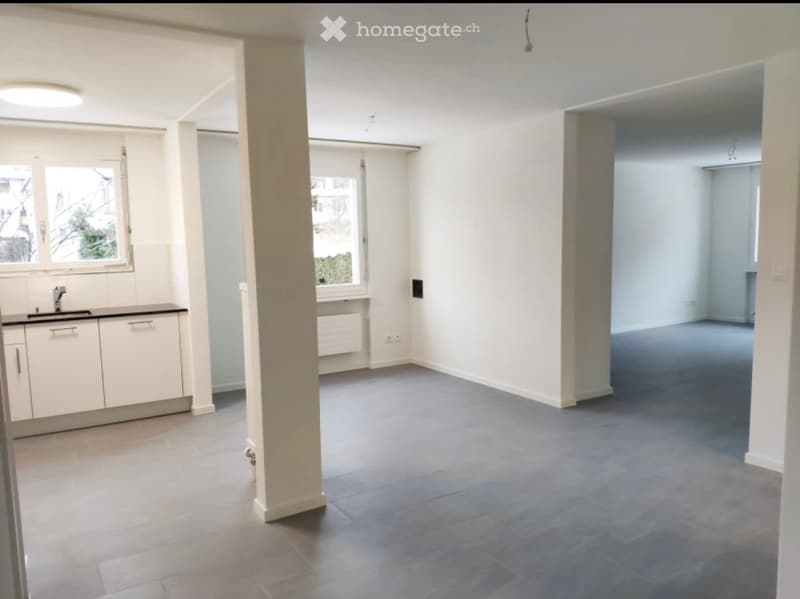 Open-plan space with grey tiled floor, white walls, central column, and a small kitchen area with a sink and cabinets.