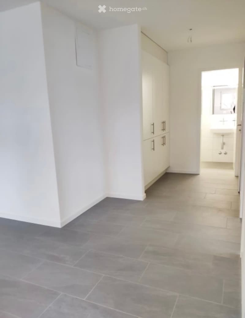 Empty room with grey tiled floor, white walls, and built-in cabinets. A doorway leads to a small bathroom area with a sink.