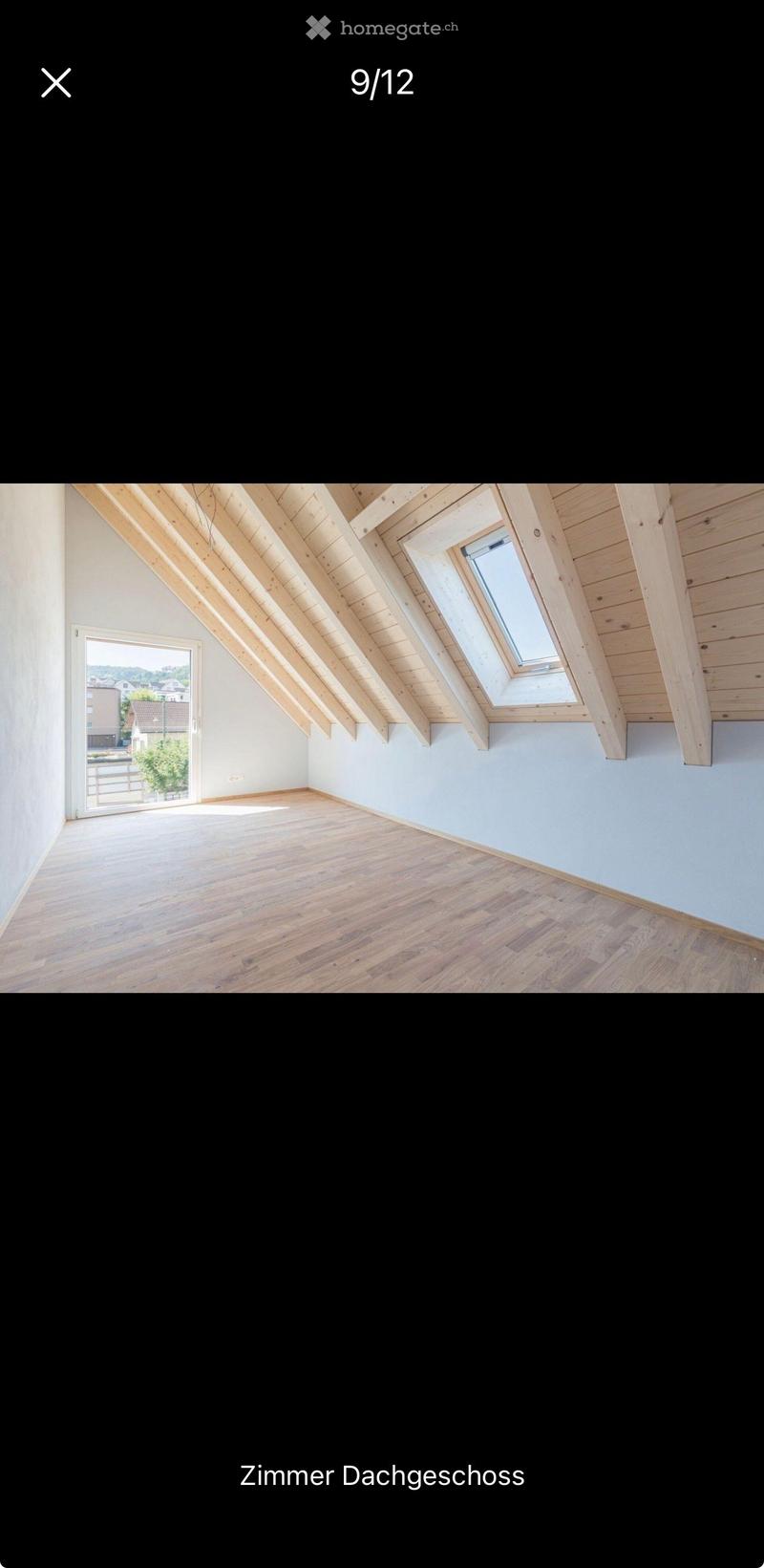 Empty attic room with wooden floors, exposed wooden beams, and skylights, featuring white walls and a large window offering a view of the outside.